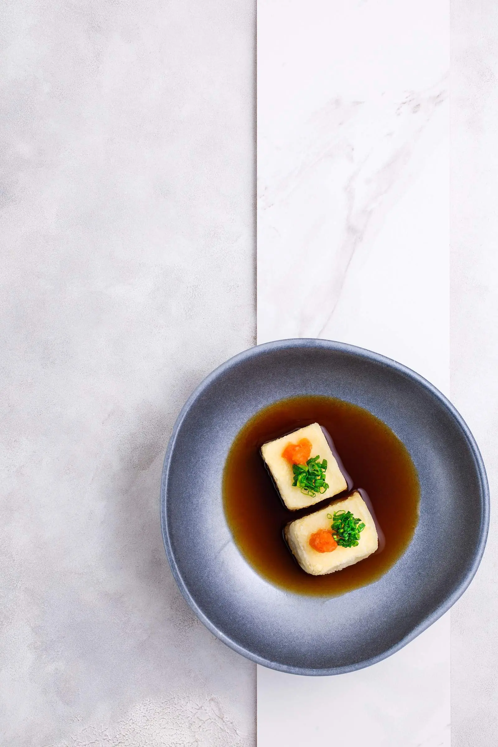 A bowl of tofu in a sauce on a marble table.